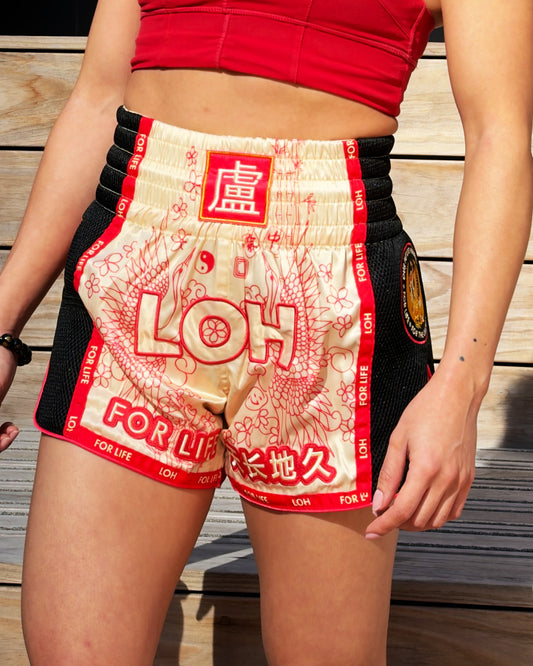 A close-up of a person wearing vintage Asian style Muay Thai shorts with a cream and red embroidered dragon crane bird irezumi tattoo style design, black mesh sides, and red trim, standing against a wooden plank background.