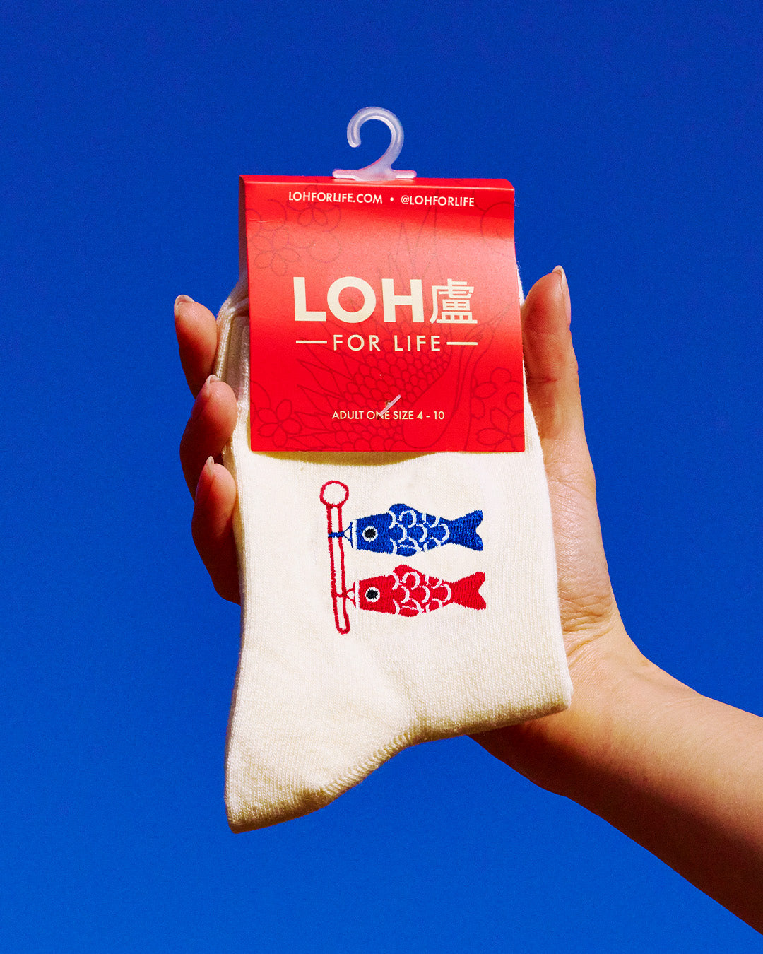 A hand holds a pair of off-white crew socks with a red and blue koi fish design and a red hanger card labeled 'LOH FOR LIFE' against a solid blue background.