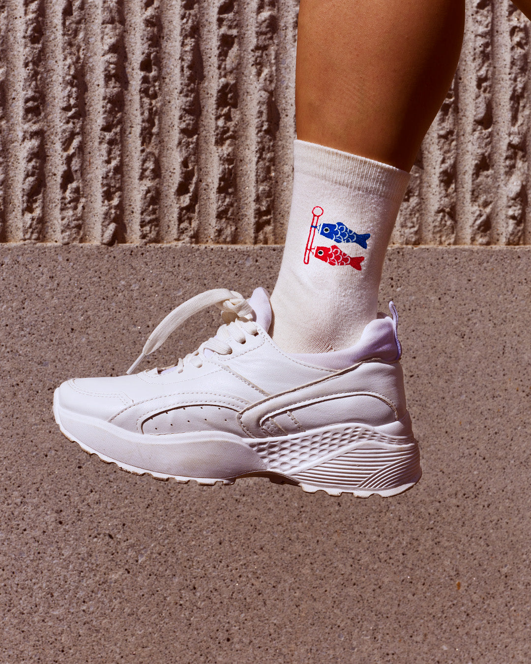 A white chunky sneaker worn with a white crew sock featuring a red and blue fish graphic, set against a textured concrete background.