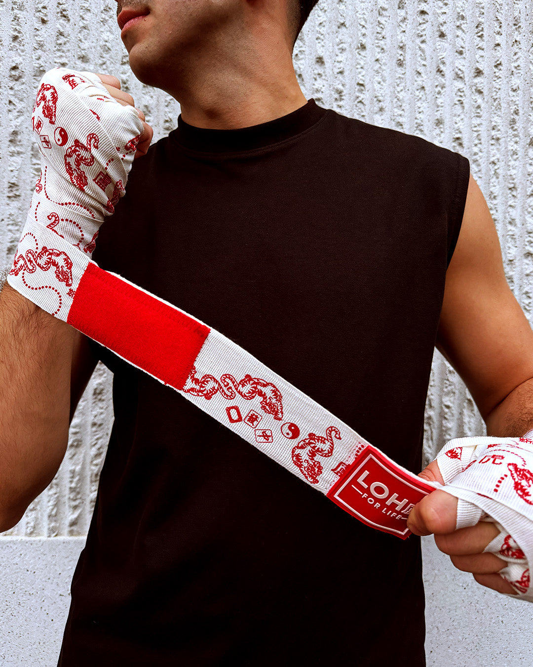 A close-up of a person wearing white boxing hand wraps with Asian inspired tiger and mahjong red graphics, holding the wrap in place against a textured light gray wall.