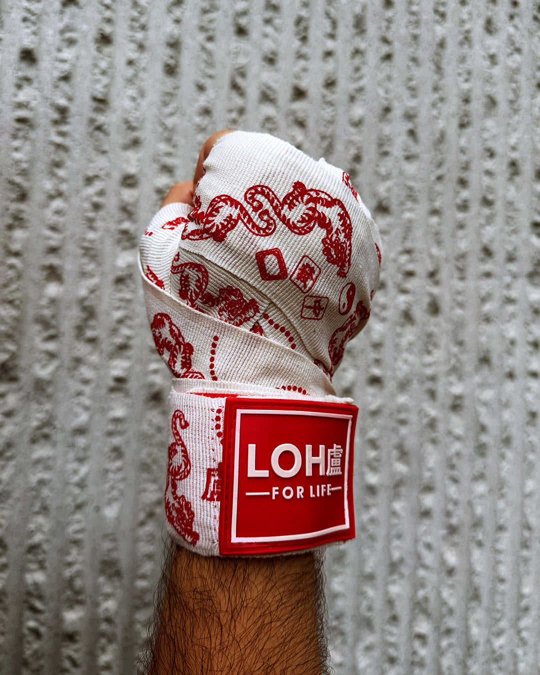 A close-up of a hand wrapped in white boxing hand wraps with red printed Asian inspired tiger mahjong graphics and a red-and-white LOH brand patch, set against a textured gray background.