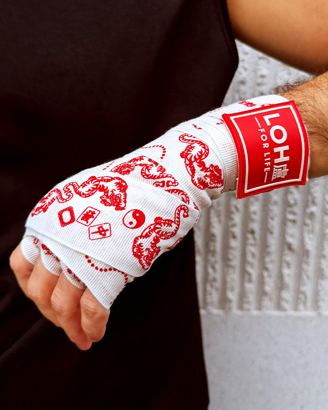 A close-up of a person's hand wrapped in white boxing wraps with red tiger, mahjong and and yin-yang patterns, with rubber label 'LOH FOR LIFE'.