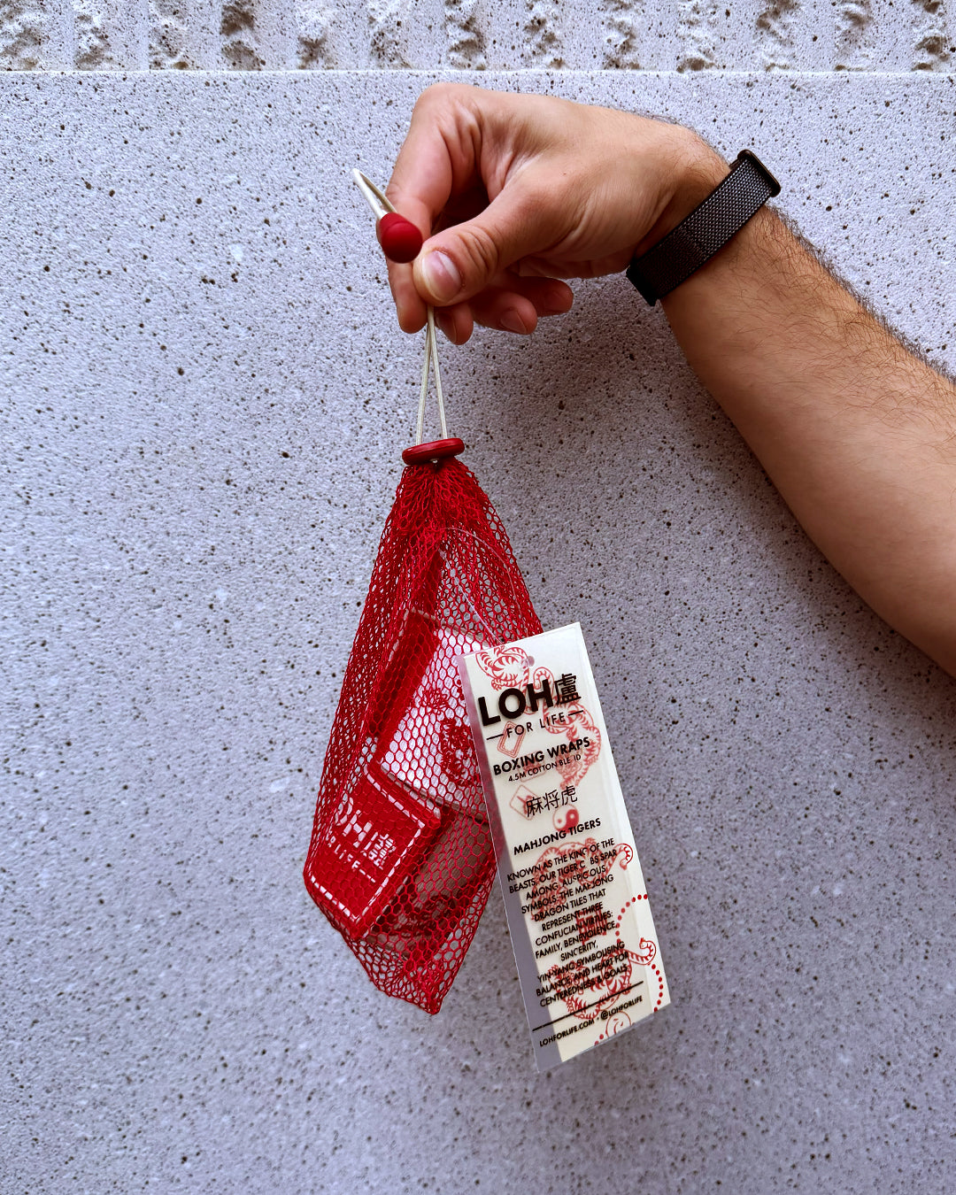 A hand holding a red mesh pouch laundry bag containing red-and-white boxing wraps against a textured light gray wall. The tag shows the brand LOH and product text for a hand care item.
