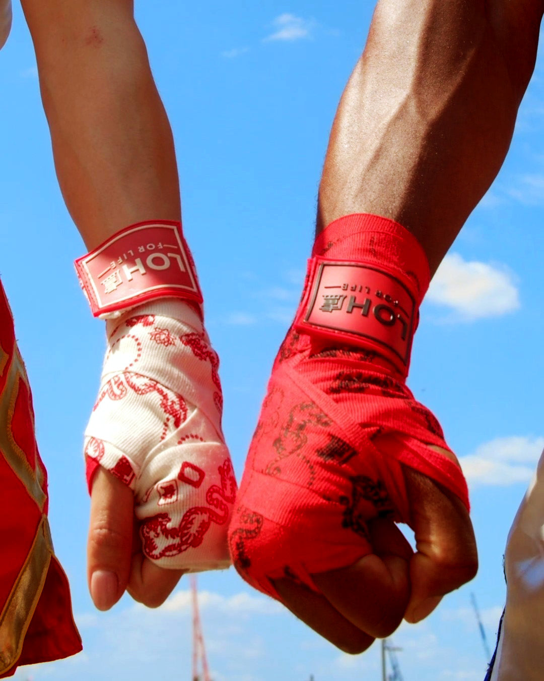 Close-up of two athletic wrists wrapped in hand wraps under a blue sky. One wrist is wrapped in white boxing wraps with red printed tiger mahjong tile graphics, and the other is wrapped in bright red fabric with a black printed details.