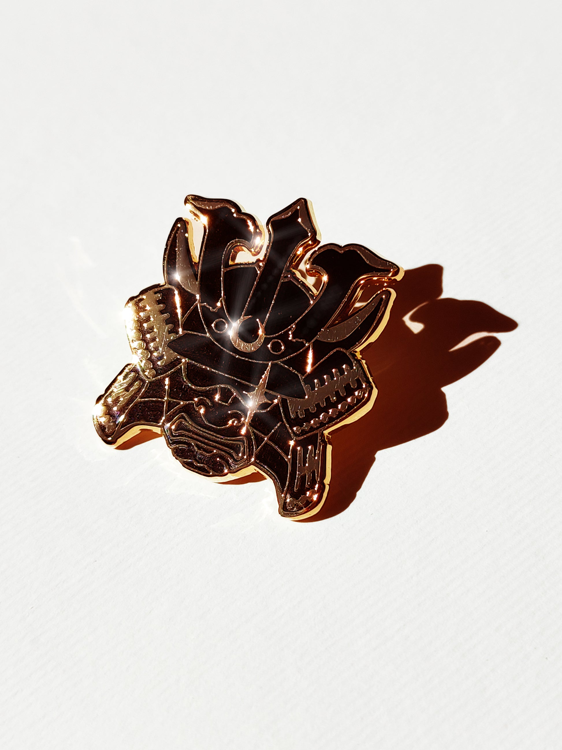 A metallic gold and black enamel pin shaped like a samurai with crescent moon and star details, photographed on a white textured background with a strong shadow.