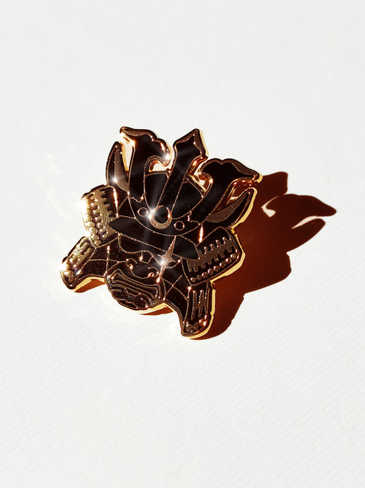A metallic gold and black enamel pin shaped like a samurai with crescent moon and star details, photographed on a white textured background with a strong shadow.
