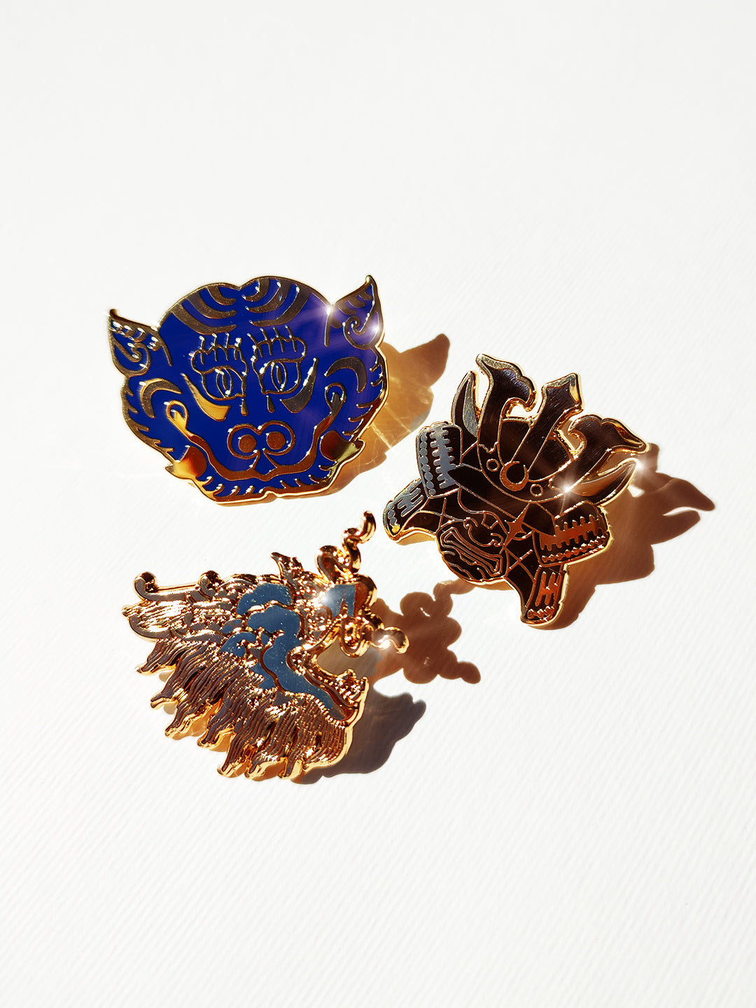 Three ornate enamel pins with gold-tone metal and blue and black enamel details, arranged on a white textured background with strong sunlight and shadows.