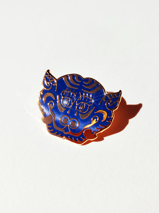 A small decorative enamel pin shaped like a stylized tiger cat head, featuring a deep cobalt blue background with gold ornamental patterns and a gold border, photographed on a plain white surface with a cast shadow.